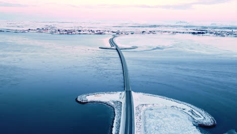 Island,-Schneebedeckte-Winterlandschaft-Mit-Straßenbrücke-über-Die-Insel-Bei-Sonnenuntergang,-Luftaufnahmen