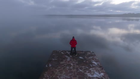 Hombre-Parado-Solo-En-Un-Muelle,-Turismo-De-Nubes-Reflejadas-En-La-Perspectiva-Aérea-De-Drones-De-Agua-De-Mar-En-La-Costa-Costera-Brumosa