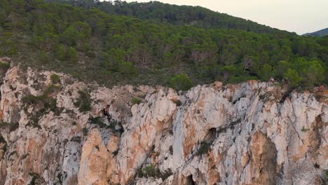 Hikers-rest-on-a-rocky-cliff-among-trees-with-a-view-of-the-green-landscape-during-sunset-in-ibiza