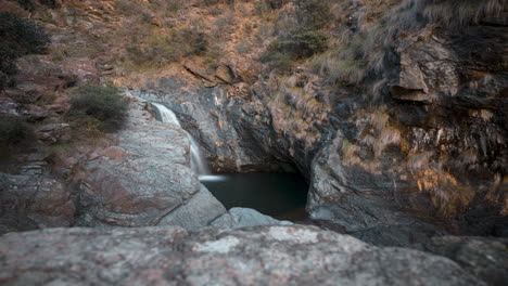 Goldener-Zeitraffer-Eines-Ruhigen-Wasserfalls,-Der-In-Einen-Felsigen-Pool-Stürzt,-Der-Von-Schroffen-Klippen-Umgeben-Ist