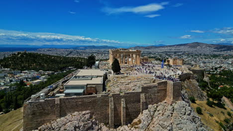 Athens,-Greece,-Aerial-View-of-Acropolis-Temple-and-Crowd-of-People-Under-Greek-National-Flag-on-Sunny-Day