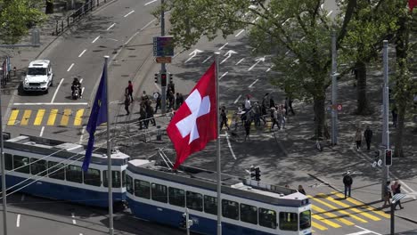 Slow-right-moving-drone-shot-of-tram-interchange-in-Zurich-with-Swiss-and-EU-flags-in-the-foreground