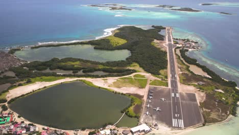Gran-roque's-airstrip-surrounded-by-turquoise-waters-and-greenery,-sunny-day,-aerial-view