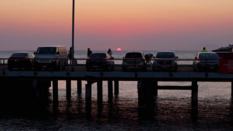 Silhouette-of-cars-parked-on-dock-pier-as-people-take-pictures-of-sun-set-below-horizon-by-the-ocean