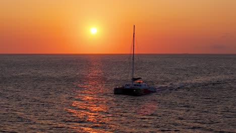 Katamaran-Fährt-Entlang-Der-Küste,-Während-Der-Sonnenuntergang-Den-Himmel-Rot-orange-Leuchten-Lässt