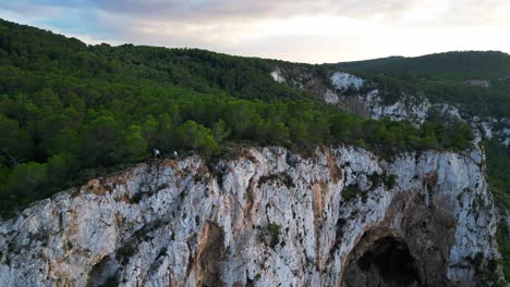 Hikers-rest-on-a-rocky-cliff-among-trees-with-a-view-of-the-green-landscape-during-sunset-in-ibiza