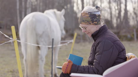 Woman-journals-with-white-horse-in-background,-equine-assisted-therapy-workshop