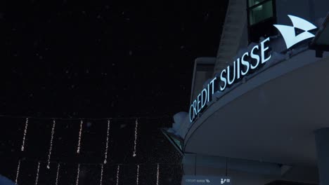 High-angle-downward-panning-shot-showing-Davos-Credit-Suisse-branch-during-the-World-Economic-Forum,-with-people-walking-past-the-camera-during-a-snowy-night