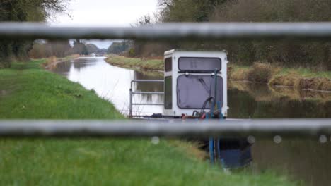 Narrowboat-cruising-on-the-Grand-Canal-in-County-Kildare,-Ireland-on-an-overcast-day,-viewed-through-grass