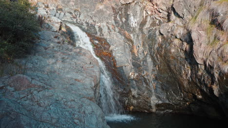 Kleiner-Wasserfall,-Der-Von-Einer-Felsklippe-In-Einen-Ruhigen-Teich-Stürzt,-Sonnenlicht-Unterstreicht-Die-Ruhige-Landschaft