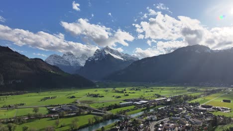An-aerial-of-the-picturesque-valley-of-Glarus-Nord,-Switzerland,-revealing-an-idyllic-scene-of-a-small-settlement-nestled-amidst-snow-capped-mountains,-serene-alpine-living-amidst-nature's-grandeur