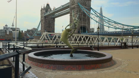 Slow-motion-tilt-up-of-Girl-with-Dolphin-statue-on-the-river-Thames,-London-with-Tower-Bridge-in-the-background