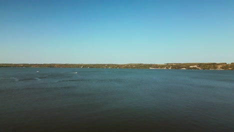 Drone-Takes-Off-Flying-Across-Large-Lake-Body-of-Water-with-Boat-and-Distant-Shore