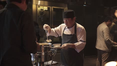 Chef-stirs-pot-in-bustling-restaurant-kitchen,-guests-visible,-warm-ambient-lighting,-close-up
