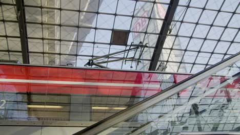 Modern-glass-train-station-with-moving-train-in-the-background,-sunlight-filtering-through