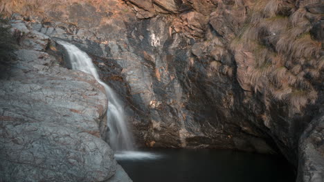 Majestuosa-Cascada-Que-Cae-En-Una-Serena-Piscina-De-Roca,-Una-Luz-Suave-Que-Acaricia-El-Terreno-Accidentado,-Vista-Aérea