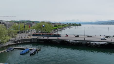 Langsame-Vorwärtsbewegung,-Schwenkende-Drohnenaufnahme-Mit-Hauptbrücke-Der-Stadt,-Schweizer-Und-EU-Flaggen,-Fußgängern,-Transportmitteln-Und-Dem-Zürichsee