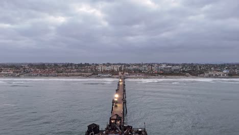 Muelle-De-Oceanside-California-Incendio-Restaurante-Dañado-Drone-Caída-Vertical-En-Revelar-Océano-Tranquilo-Amanecer-Nublado-Con-La-Costa-En-El-Fondo