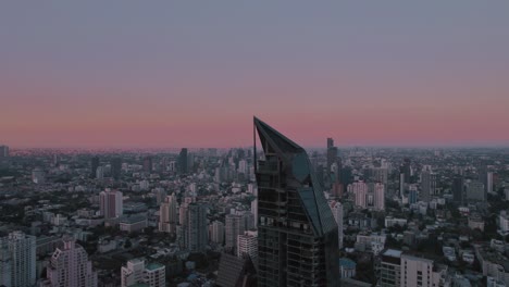 Aerial-vantage-point,-Bangkok's-skyline-unfolds,-displaying-urban-life-against-a-backdrop-where-the-horizon-takes-on-post-sunset-hues-of-red-and-violet