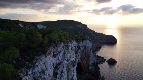 Hikers-rest-on-a-rocky-cliff-among-trees-with-a-view-of-the-green-landscape-during-sunset-in-ibiza