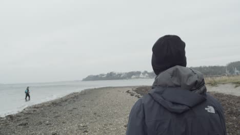 Guy-walks-on-beach-next-to-water-on-windy-and-cloudy-day,-tracking-shot,-slow-motion