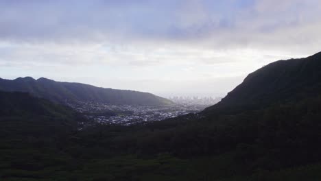 Drone-footage-passing-between-two-lush-green-mountains-to-reveal-a-town-between-then-with-a-silhouetted-view-of-the-city-Honolulu-on-the-horizon-in-the-mist