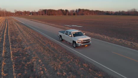 Schwenkaufnahme-Eines-Pickups,-Der-Von-Hinten-Auf-Einer-Landstraße-Auf-Einem-Bauernhof-In-Den-Sonnenuntergang-Fährt