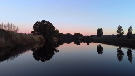 Aerial-drone-view-flying-low-in-the-centre-of-a-wide-river-at-twilight-with-trees-and-grass-on-river-bank