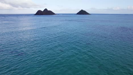 Imágenes-De-Drones-En-El-Océano-Pacífico-Cerca-De-Oahu,-Hawaii,-Que-Muestran-La-Refracción-De-Las-Olas-Cuando-Las-Olas-Del-Océano-Son-Interrumpidas-Por-Los-Islotes-Y-El-Agua-Azul-Verde.