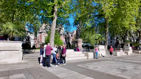 Tour-group-gathers-around-Mahatma-Gandhi-statue-in-Parliament-Square-on-a-sunny-day,-green-trees