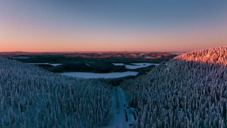 Aerial-view-over-a-road-in-middle-of-forested-hills,-winter-evening-in-Lapland