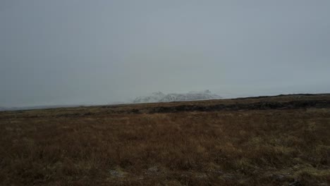 Low-Aerial-Drone-Shot-Over-Iceland's-Landscape-with-Grass-and-Wintery-Mountainous-Background