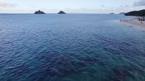 Imágenes-De-Drones-A-Través-Del-Océano-Pacífico-Cerca-De-Oahu-Hawaii-Con-Una-Playa-De-Arena-Blanca-A-La-Derecha-Y-Dos-Pequeños-Islotes-En-El-Horizonte-Y-Arrecifes-De-Coral-En-El-Fondo-Del-Océano