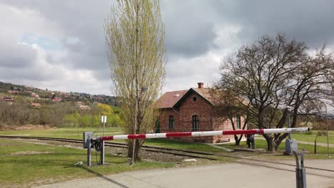 Kleiner-Zug,-Der-An-Einem-Bewölkten-Tag-An-Einem-Kleinen-Bahnhofsgebäude-Mit-Einer-Eisenbahnschranke-Auf-Dem-Land-Vorbeifährt