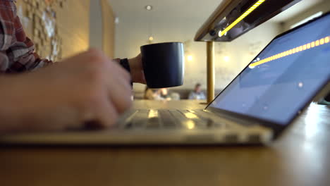 Cerca-De-Las-Manos-De-Un-Hombre-Escribiendo-En-Un-Cuaderno-Y-Tomando-Café,-Dentro-De-Un-Acogedor-Bar-Con-Gente-Al-Fondo