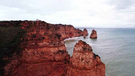 Coastline-establishing-shot-with-orange-rugged-cliffs-and-sea-stacks-by-aerial-4k-drone-at-Ponta-da-Piedade-near-Lagos-in-the-Algarve-region-of-Portugal