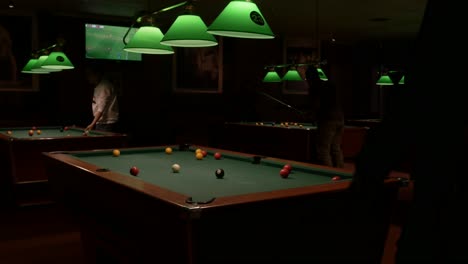 People-playing-on-pool-tables-at-a-pool-hall-at-night-with-sports-on-television-playing-in-the-background