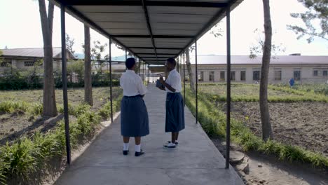 Two-African-Girls-in-uniform-heading-to-class-in-a-private-school,-walking-underneath-a-roof-surrounded-by-a-garden-with-trees-and-plants