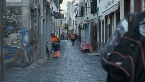 Still-shot-worker-cleaning-cobbled-street-with-tourists-passing-by,-Funchal-old-town