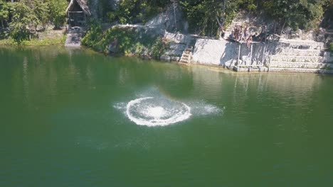 Aerial-drone-shot-of-a-man-jumping-from-a-rope-doing-a-backflip-into-the-lake-in-flores-peten,-guatemala