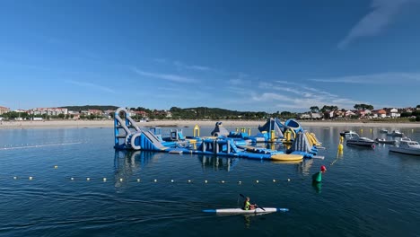 A-European-and-blonde-boy-rowing-intensely-in-his-canoe-over-to-the-water-of-the-beach-between-the-boats-with-the-castle-of-inflatable-slides-nearby,-tracking-shot