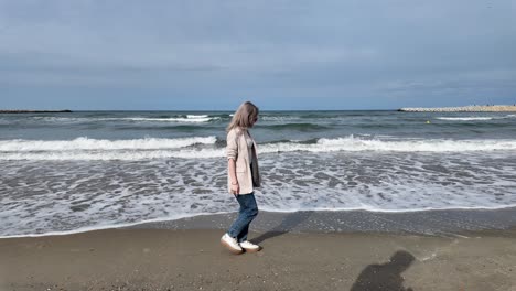 A-Slow-Motion-Shot-of-a-Blonde-Girl-Walking-on-the-Beach-with-Waves-Slowly-Hitting-the-Shoreline