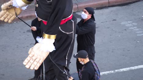 Tres-Hombres-Tirando-De-Los-Hilos-Para-Mover-Un-Títere-Gigante-En-La-Calle,-Todos-Con-Mascarilla-Protectora-Para-El-Coronavirus