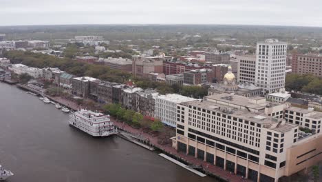 Toma-Aérea-Descendente-Y-Panorámica-De-Un-Crucero-Fluvial-Atracado-A-Lo-Largo-Del-Río-En-El-Centro-De-Savannah,-Georgia