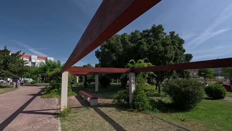 Roofless-wooden-structure-with-granite-supports-in-the-middle-of-the-public-park-where-tourists-walk-in-summer-on-a-day-of-radiant-sun-and-heat,-crane-firing-going-down,-Pontevedra,-Galicia,-Spain