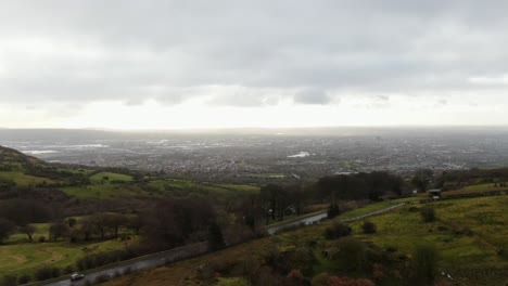 Drone-panning-right-with-fields,-car-driving-on-a-road,-farmland-and-city-in-the-background