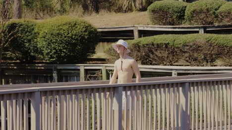 Caucasian-lean-man-with-bathers-and-sun-hat-walking-on-wooden-jetty-during-sunlight-and-enjoying-holidays-at-ocean