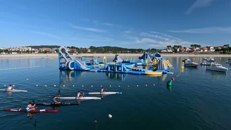 Los-Niños-Reman-En-Sus-Canoas-Ante-El-Enorme-Castillo-Hinchable-De-Desafíos-Físicos-Y-Toboganes-Flotando-En-El-Agua-Con-La-Playa-Al-Fondo,-Toma-De-Seguimiento.