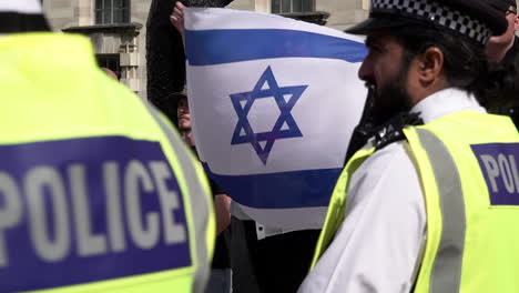 Two-Metropolitan-police-officers-wearing-yellow-tabards-stand-and-monitor-a-public-order-situation-as-a-person-waves-a-Star-of-David-Israeli-flag