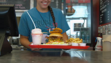 Woman-employee-at-a-burger-joint-pushes-a-full-tray-with-the-largest-burger-and-fries-and-shake-across-the-counter-for-the-customer-to-pick-up